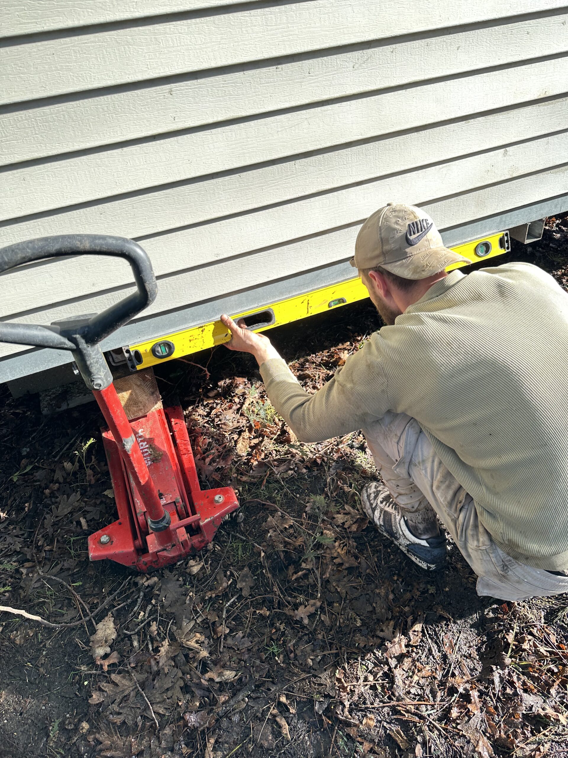 Ouvrier utilisant un niveau à bulle jaune et un cric hydraulique rouge pour niveler une maison mobile ou un abri de jardin.